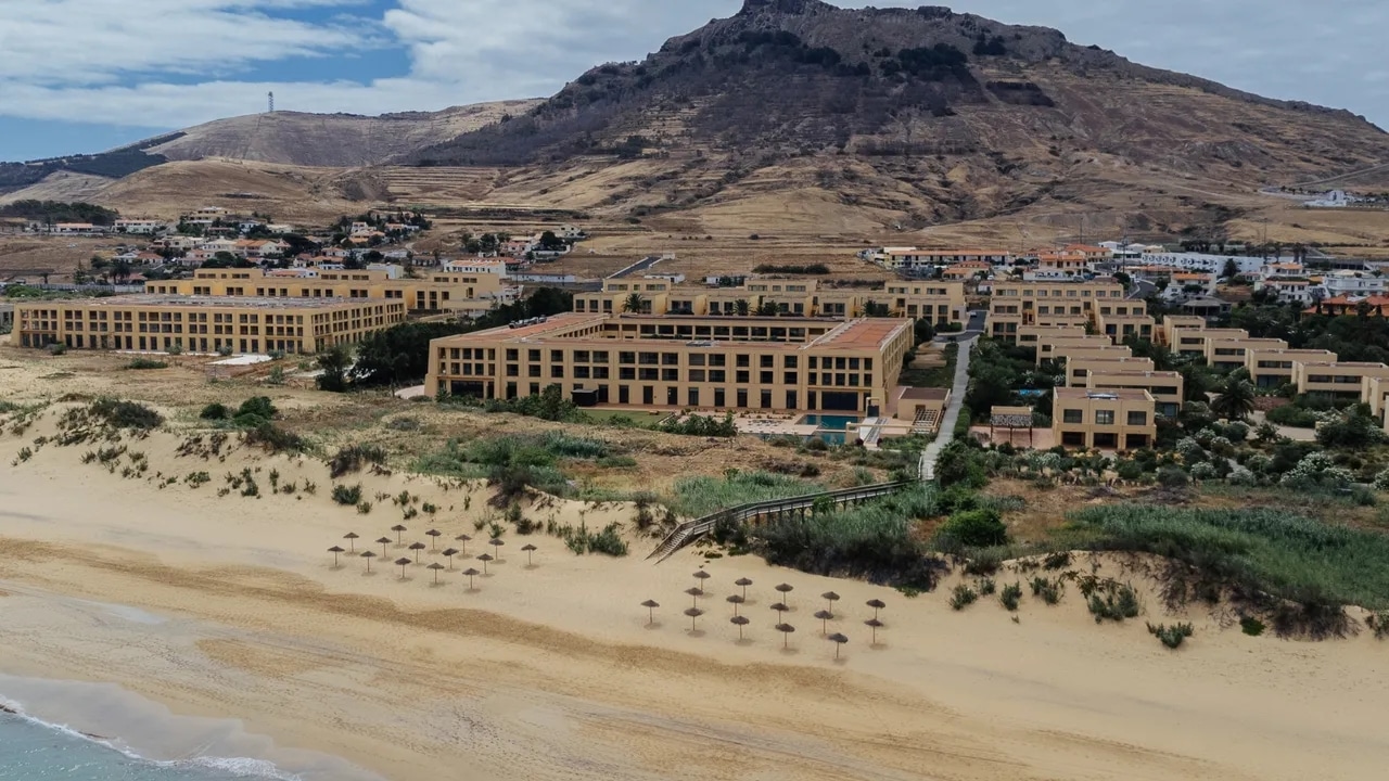 http://Vue%20aérienne%20d’un%20hôtel%20en%20bord%20de%20plage%20à%20Porto%20Santo,%20entre%20dunes%20de%20sable%20et%20reliefs%20montagneux.