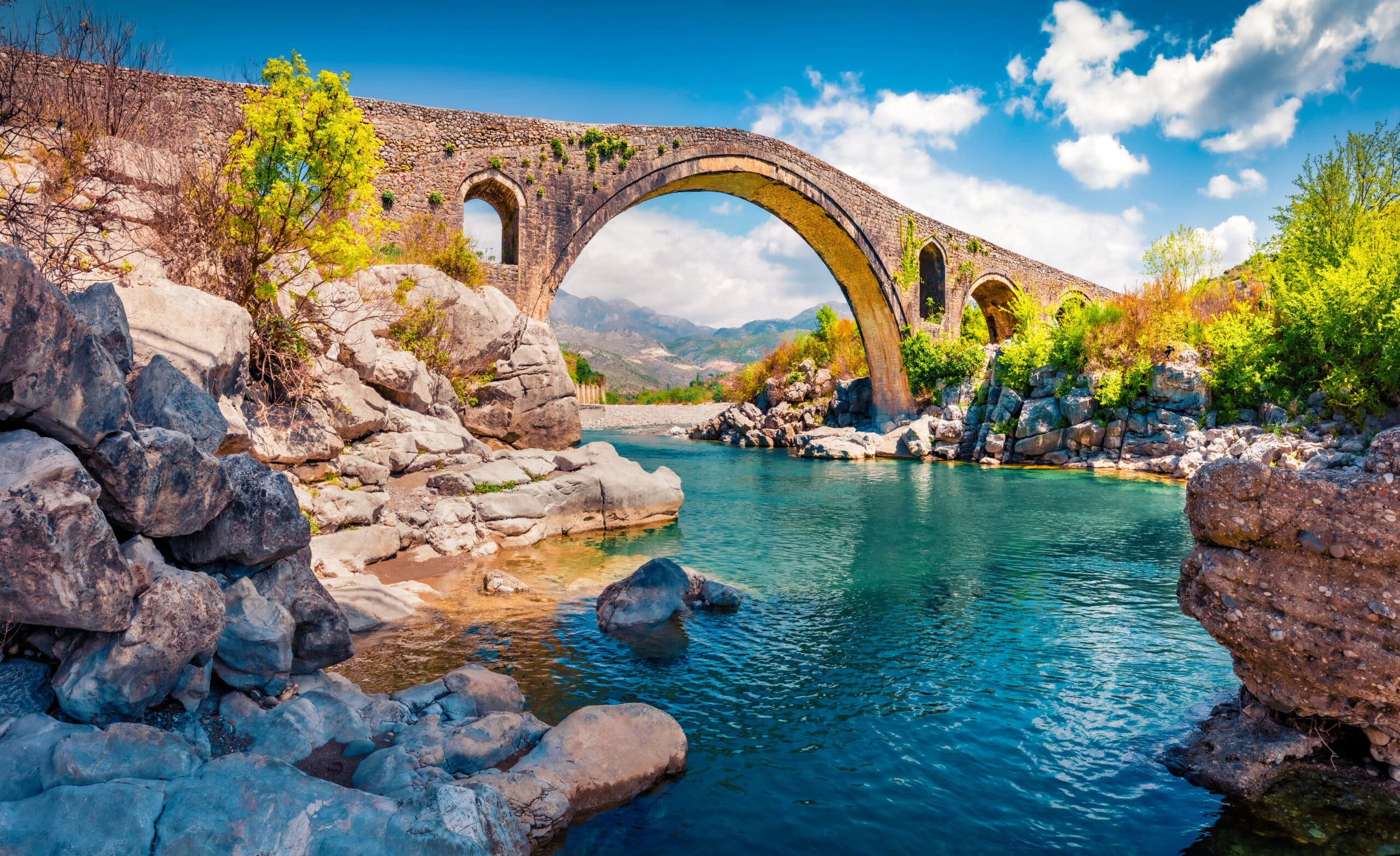 Pont historique en pierre à arches franchissant une rivière turquoise dans les montagnes albanaises.