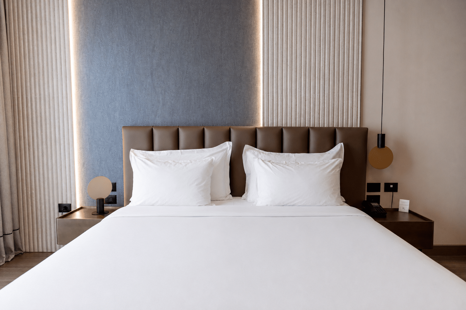 Chambre d’hôtel moderne en Albanie avec grand lit double, tête de lit capitonnée et éclairage contemporain.