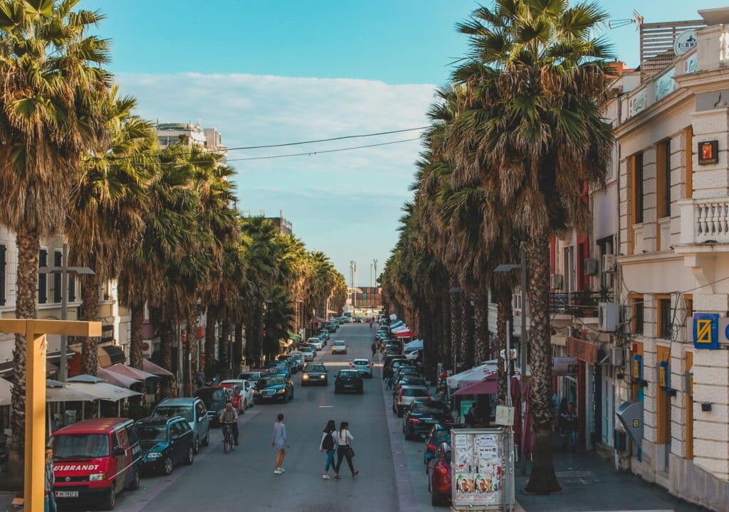 Rue animée d’une ville côtière d’Albanie bordée de palmiers, commerces et terrasses.