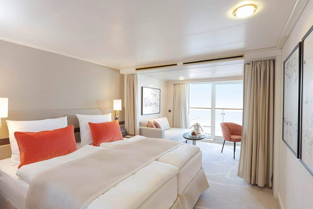 Cabine lumineuse de bateau de croisière avec lit double, coin salon et balcon donnant sur la mer.