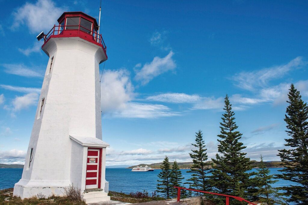 Phare blanc et rouge sur la côte canadienne avec un bateau de croisière naviguant au large.
