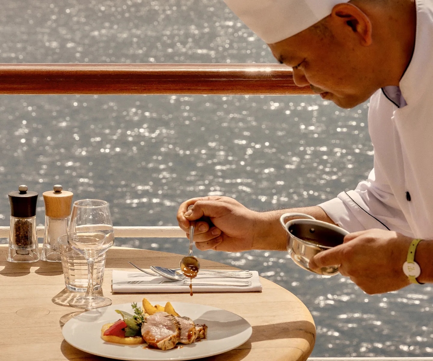 Chef cuisinier en tenue blanche dressant une assiette gastronomique avec une sauce sur une table de restaurant en plein air face à la mer.
